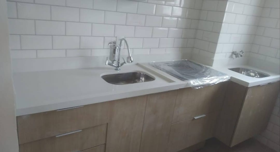 Cozinha com bancada branca em L, duas cubas de inox e recorte para cooktop