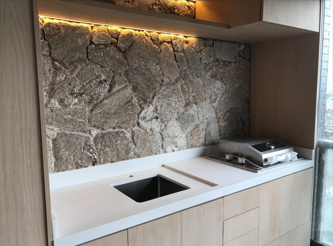 Espaço gourmet com bancada branca, cuba quadrada e parede revestida em pedra rústica